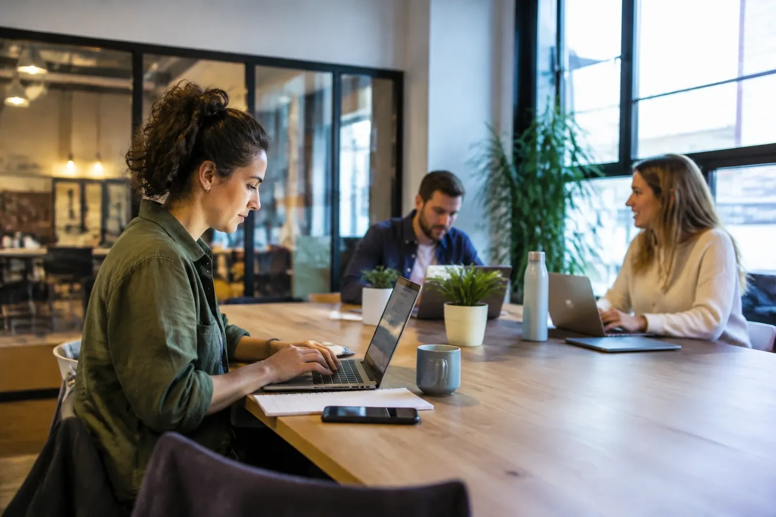Vrouw werkt geconcentreerd op laptop aan grote tafel, twee collega’s praten op achtergrond